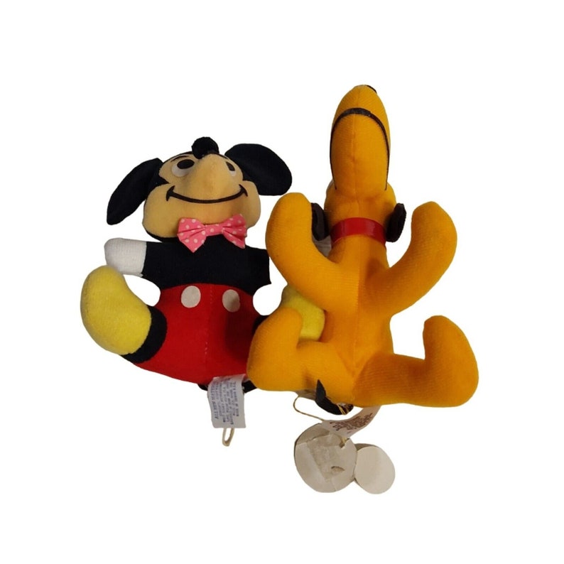 Op de afbeelding: Twee pluche Disney-personages. Mickey Mouse draagt een zwart smoking met een rode broek, gele schoenen en een roze vlinderdas. Pluto is geel met zwarte oren en een rode halsband. Op een witte achtergrond.