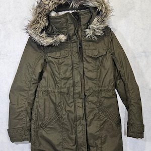 May include: An olive green parka-style jacket with a faux fur-trimmed hood. The jacket features multiple pockets, a zipper closure, and a drawstring waist. The hood has a tan and brown faux fur trim. The jacket is made of a durable, water-resistant material.