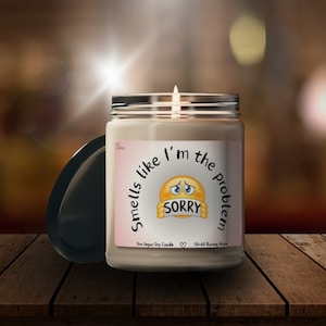 Op de afbeelding: Een sojawas kaars met een wit label waarop staat "Smells like I'm the problem" met een geel emoji-gezicht dat zijn mond bedekt met een bordje waarop "Sorry" staat. De kaars zit in een glazen pot met een zwarte deksel.