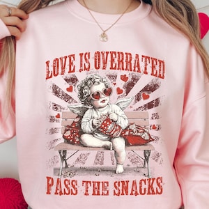 May include: A light pink sweatshirt with the text "LOVE IS OVERRATED PASS THE SNACKS". The graphic features a cherub wearing heart-shaped sunglasses, sitting on a bench and holding snacks. The design has a vintage, distressed look.