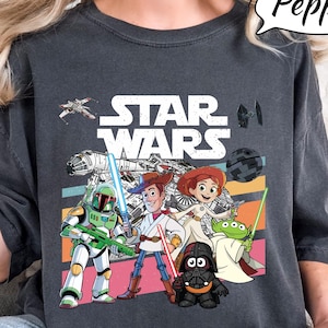 May include: Dark gray t-shirt featuring a colorful graphic of Star Wars characters, including Woody, Buzz Lightyear, and Darth Vader. The Star Wars logo is prominently displayed above the characters. The shirt has a relaxed fit.