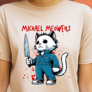 May include: Beige t-shirt featuring a cartoon cat dressed as Michael Myers, holding a knife. The text "MICHAEL MEOWERS" is printed above the cat in red. The cat is wearing a blue jumpsuit and sneakers, with red splatter details.