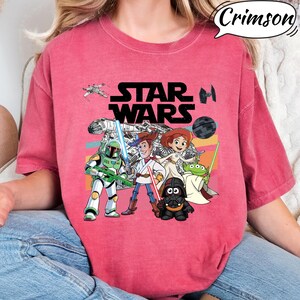 May include: A red t-shirt featuring a Star Wars graphic with characters from the movie and the text "STAR WARS". The design includes characters like Darth Vader, Woody, and Buzz Lightyear.