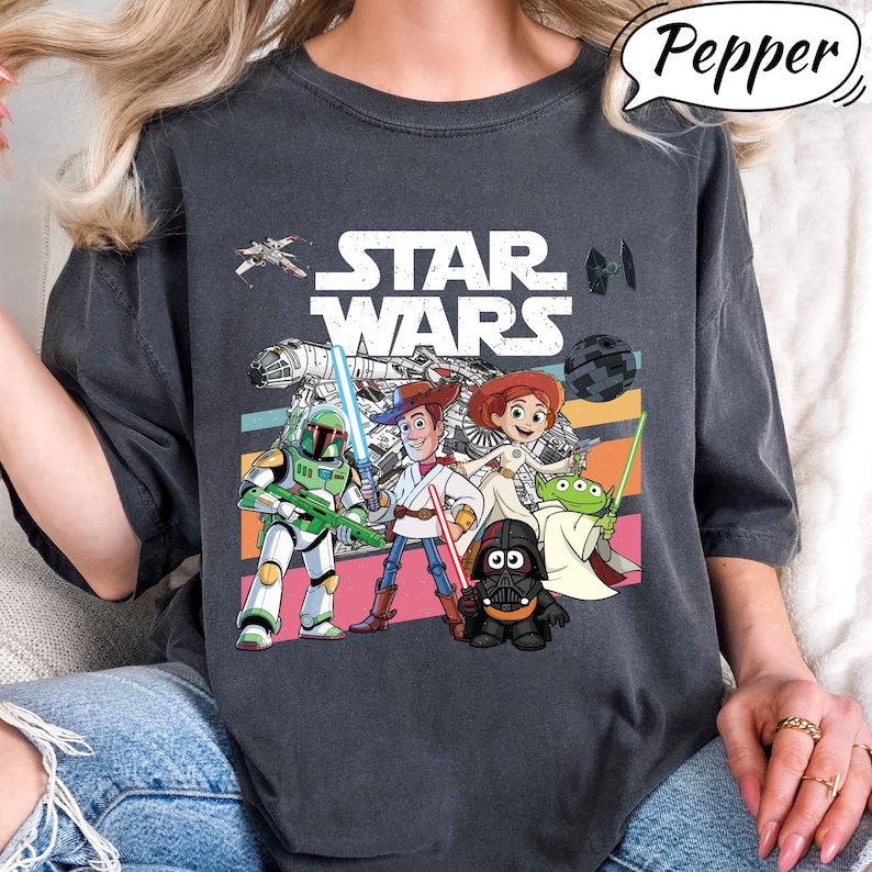 May include: Dark gray t-shirt featuring a colorful graphic of Star Wars characters, including Woody, Buzz Lightyear, and Darth Vader. The Star Wars logo is prominently displayed above the characters. The shirt has a relaxed fit.