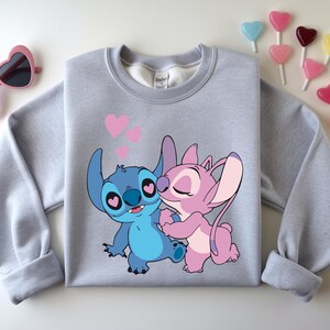 Op de afbeelding: Een lichtgrijs sweatshirt met een cartoonafbeelding van Stitch en Angel die elkaar omhelzen, met roze harten erboven. De sweatshirt heeft lange mouwen en een ronde hals. Roze hartvormige zonnebrillen en hartvormige lolly's staan ook op de afbeelding.