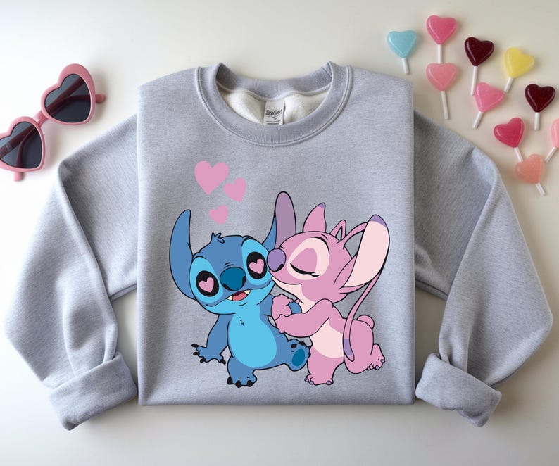 Op de afbeelding: Een lichtgrijs sweatshirt met een cartoonafbeelding van Stitch en Angel die elkaar omhelzen, met roze harten erboven. De sweatshirt heeft lange mouwen en een ronde hals. Roze hartvormige zonnebrillen en hartvormige lolly's staan ook op de afbeelding.