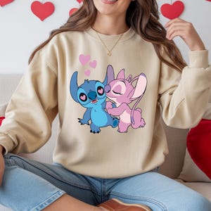 Op de afbeelding: Een beige sweatshirt met een cartoonillustratie van Stitch en Angel die elkaar omarmen, met roze hartjes. De personages zijn blauw en roze. Het sweatshirt wordt gedragen door een persoon in een blauwe spijkerbroek.