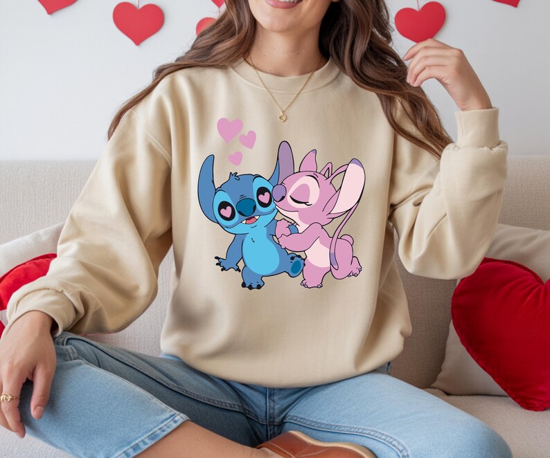 Op de afbeelding: Een beige sweatshirt met een cartoonillustratie van Stitch en Angel die elkaar omarmen, met roze hartjes. De personages zijn blauw en roze. Het sweatshirt wordt gedragen door een persoon in een blauwe spijkerbroek.