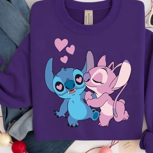 Op de afbeelding: Een paarse sweatshirt met een afbeelding van twee stripfiguren, een blauwe en een roze, die elkaar omarmen. Roze harten zweven erboven. De sweatshirt heeft lange mouwen en een label bij de hals.