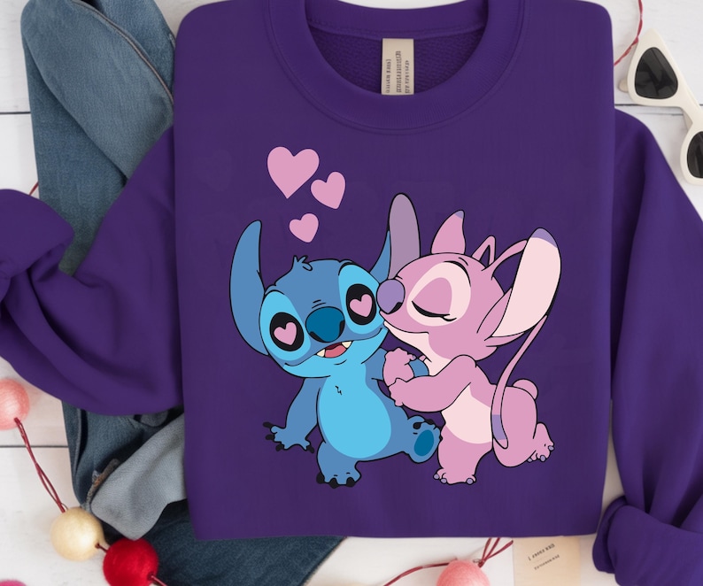 Op de afbeelding: Een paarse sweatshirt met een afbeelding van twee stripfiguren, een blauwe en een roze, die elkaar omarmen. Roze harten zweven erboven. De sweatshirt heeft lange mouwen en een label bij de hals.