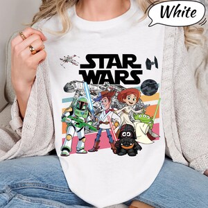 May include: White t-shirt featuring a colorful graphic of Star Wars characters, including Darth Vader, Yoda, and Woody. The Star Wars logo is prominently displayed above the characters. The shirt has a retro design with horizontal stripes.