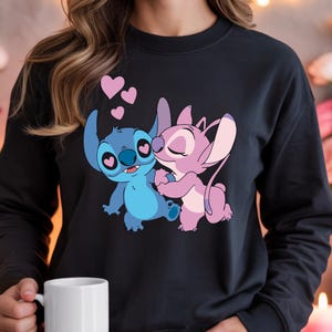 Op de afbeelding: Zwart sweatshirt met een cartoonillustratie van Stitch en Angel die elkaar omhelzen, met roze harten erboven. De personages zijn in blauw- en roze tinten. Het sweatshirt heeft een ronde hals.