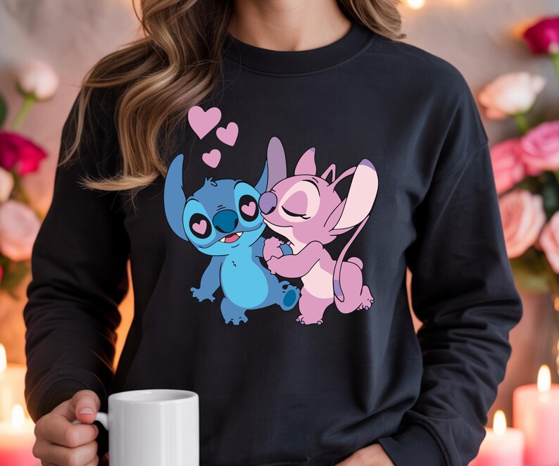 Op de afbeelding: Zwart sweatshirt met een cartoonillustratie van Stitch en Angel die elkaar omhelzen, met roze harten erboven. De personages zijn in blauw- en roze tinten. Het sweatshirt heeft een ronde hals.
