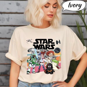 May include: Ivory-colored t-shirt featuring a Star Wars graphic with cartoon characters. The design includes the Star Wars logo, a spaceship, and various characters from the franchise. The shirt has short sleeves and a relaxed fit.