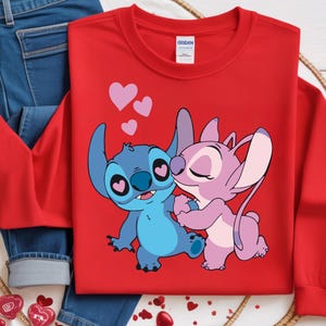 Op de afbeelding: Een rode sweatshirt met een cartoonillustratie van twee personages, &eacute;&eacute;n blauw en &eacute;&eacute;n roze, die elkaar omarmen. Het blauwe personage heeft hartvormige ogen en het roze personage kust het blauwe. Roze harten zweven boven hen.