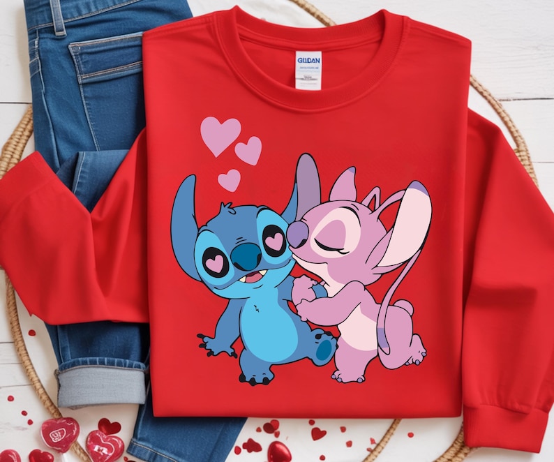 Op de afbeelding: Een rode sweatshirt met een cartoonillustratie van twee personages, &eacute;&eacute;n blauw en &eacute;&eacute;n roze, die elkaar omarmen. Het blauwe personage heeft hartvormige ogen en het roze personage kust het blauwe. Roze harten zweven boven hen.