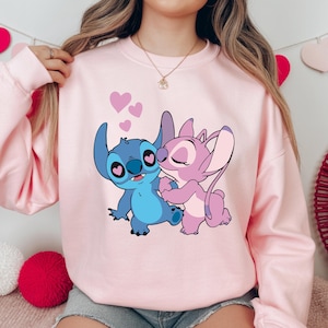 Op de afbeelding: Een lichtroze sweatshirt met een cartoonillustratie van Stitch en Angel, met harten eromheen. De personages zijn in blauw- en roze tinten, omarmend. De sweatshirt heeft lange mouwen en een ronde hals.