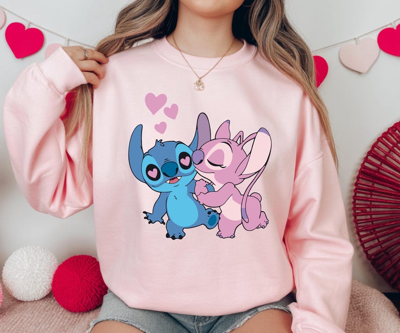 Op de afbeelding: Een lichtroze sweatshirt met een cartoonillustratie van Stitch en Angel, met harten eromheen. De personages zijn in blauw- en roze tinten, omarmend. De sweatshirt heeft lange mouwen en een ronde hals.