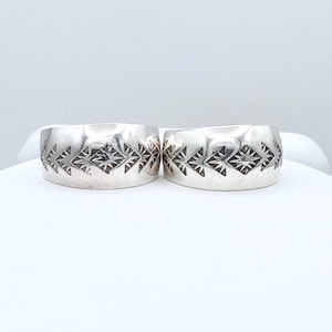 May include: Two wide silver rings featuring a polished finish and a central band of stamped geometric patterns. Each ring displays a repeating sequence of triangular shapes and a star-like motif engraved into the metal surface, creating a textured design.