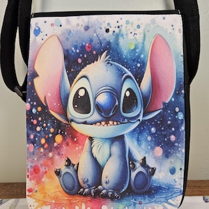 May include: A small, square crossbody bag featuring a watercolor illustration of the Disney character Stitch. The bag has a black strap and trim, with a colorful background of blues, pinks, and yellows. The image of Stitch is centered on the bag.