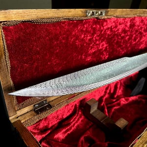 May include: A patterned steel blade is displayed in an open wooden box lined with red velvet. The blade features a distinctive wavy design and is presented on a wooden stand. The box's lid is open, showcasing the blade's full length.