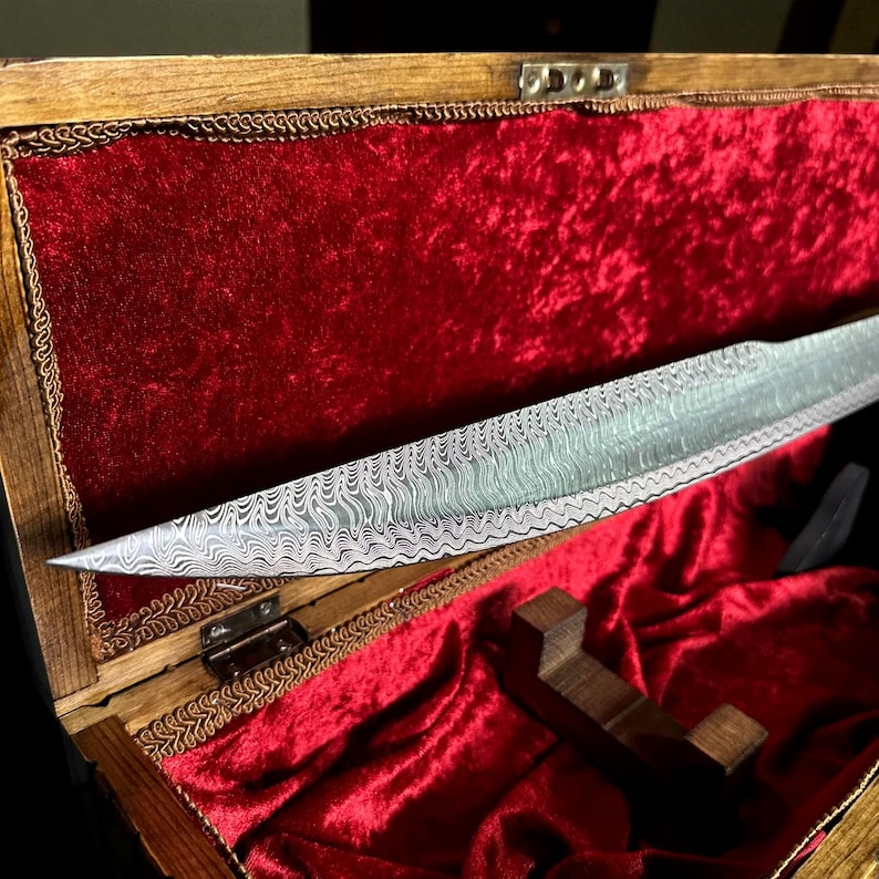 May include: A patterned steel blade is displayed in an open wooden box lined with red velvet. The blade features a distinctive wavy design and is presented on a wooden stand. The box's lid is open, showcasing the blade's full length.