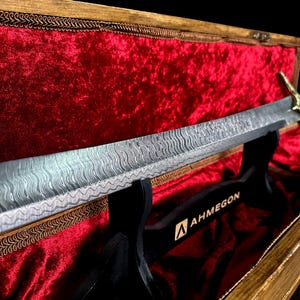 May include: A decorative sword with a patterned blade rests on a black stand inside a wooden box lined with red velvet. The sword has a gold-coloured handle and the stand has the word "AHMEGON" printed on it.