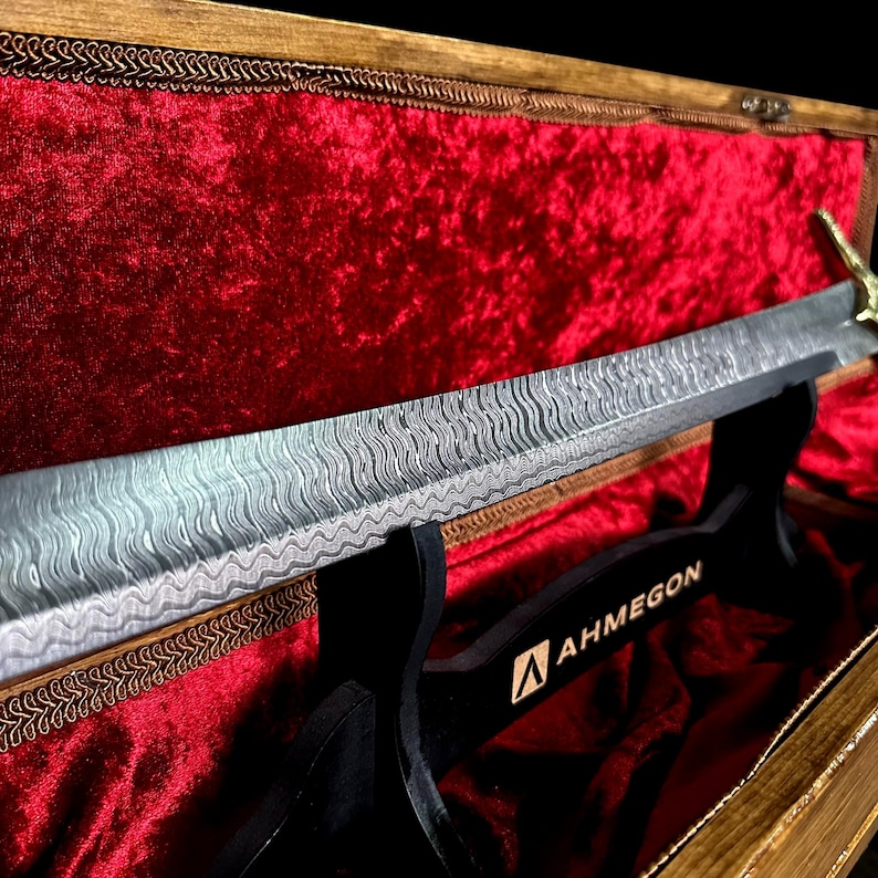 May include: A decorative sword with a patterned blade rests on a black stand inside a wooden box lined with red velvet. The sword has a gold-coloured handle and the stand has the word "AHMEGON" printed on it.