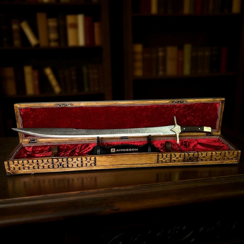 May include: A decorative sword with a silver blade and black handle, presented in an open wooden case lined with red velvet. The case features detailed carvings, with the brand name "AHMEGON" visible.