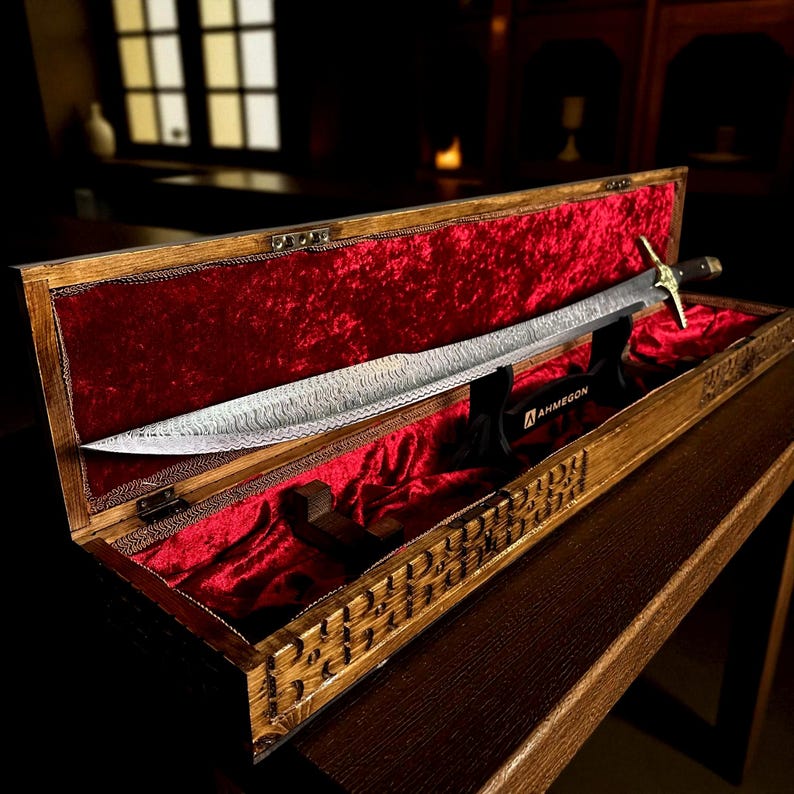May include: A decorative sword in a wooden case. The sword has a patterned blade and a gold-coloured hilt. The case is lined with red velvet and has a carved wooden exterior. The sword is displayed on a black stand.