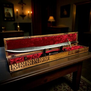 May include: A long, curved sword with a patterned blade is displayed in an open wooden case lined with red velvet. The sword's hilt is visible, along with the "AHMEGON" logo. The wooden case features engraved decorative patterns.