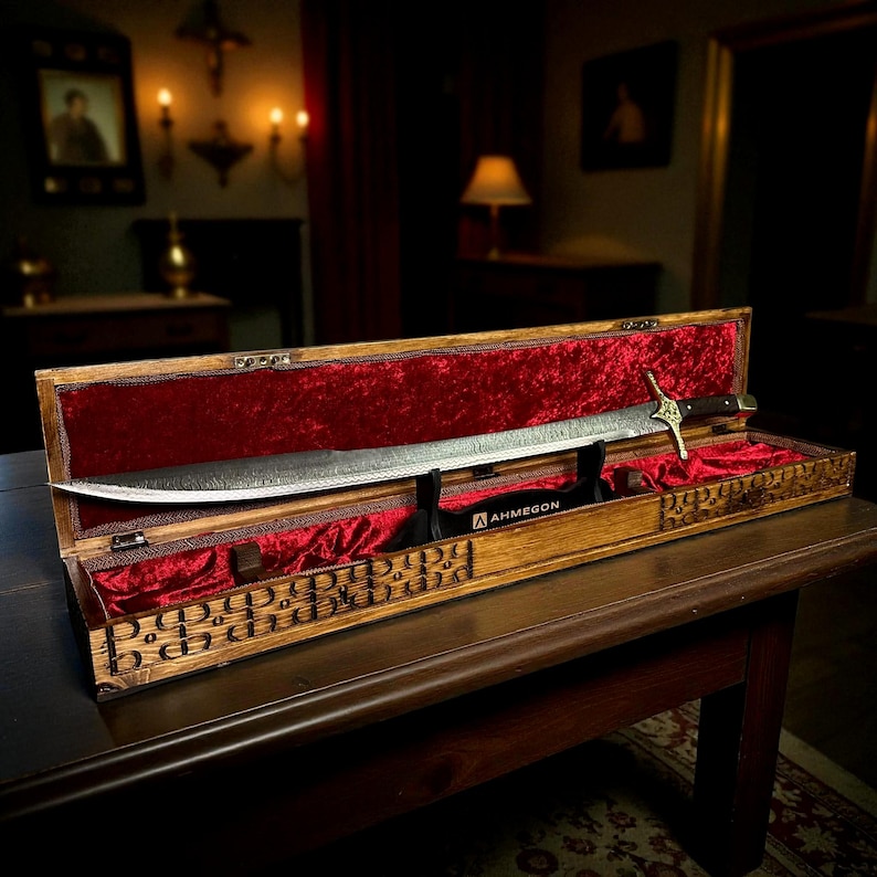 May include: A long, curved sword with a patterned blade is displayed in an open wooden case lined with red velvet. The sword's hilt is visible, along with the "AHMEGON" logo. The wooden case features engraved decorative patterns.