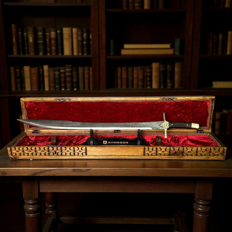 May include: A long, silver sword with a decorative gold and black handle is displayed in an open wooden case lined with red velvet. The case sits on a wooden table, with a bookshelf in the background. The sword's blade is visible.