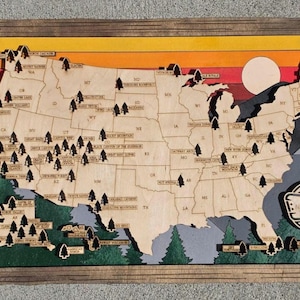 May include: A wooden wall map of the United States featuring laser-cut state borders and small tree icons marking national parks. The background displays a sunset gradient of yellow, orange, and red, with a white sun and dark green pine trees.