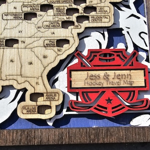 Pu&ograve; includere: Una mappa in legno degli Stati Uniti con gli stadi di hockey segnati. La mappa &egrave; incorniciata in legno scuro e presenta un design di bastone da hockey rosso e bianco con il testo "Jess & Jenn Hockey Travel Map" al centro.