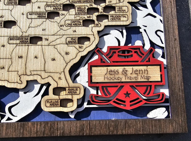 Pu&ograve; includere: Una mappa in legno degli Stati Uniti con gli stadi di hockey segnati. La mappa &egrave; incorniciata in legno scuro e presenta un design di bastone da hockey rosso e bianco con il testo "Jess & Jenn Hockey Travel Map" al centro.