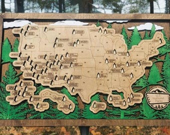 May include: A wooden map of the United States with all of the national parks marked with small pine tree icons. The map is surrounded by a wooden frame and green pine trees. The text "U.S. National Parks" is printed on the bottom right corner of the map.