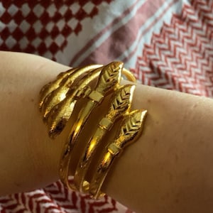 Puede incluir: Un conjunto de cuatro pulseras de brazalete doradas con un diseño de cabeza de serpiente. Cada pulsera tiene una superficie texturizada y un diseño de punta de flecha en relieve.