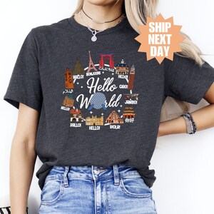 Puede incluir: Una camiseta gris oscuro con un gráfico de un globo terráqueo rodeado de varios monumentos de todo el mundo. El texto "Hello World" está impreso en blanco en el globo.