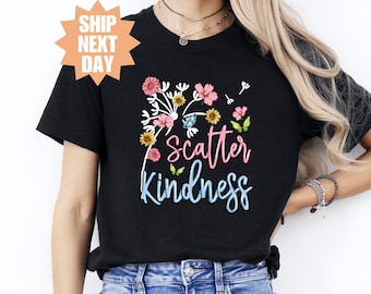 Puede incluir: Camiseta negra con un gráfico de diente de león y el texto "Scatter Kindness" en letras azules y rosas. El diente de león está rodeado de flores coloridas y mariposas.