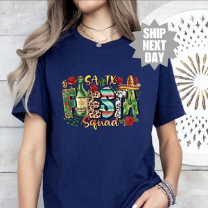 May include: A navy blue t-shirt with a graphic design that says "SA TX Fiesta Squad" with a bottle of tequila, a sombrero, and floral accents.
