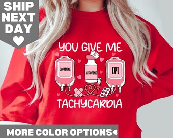 Op de afbeelding: Rood sweatshirt met een afbeelding van drie infuuszakken met de labels "Dopamine", "Atropine" en "EPI" met een hart en de tekst "You Give Me Tachycardia".