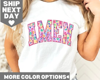 Puede incluir: Camiseta blanca con la palabra "AMEN" en letras rosas delineadas. Las letras están rellenas con un estampado de leopardo colorido.