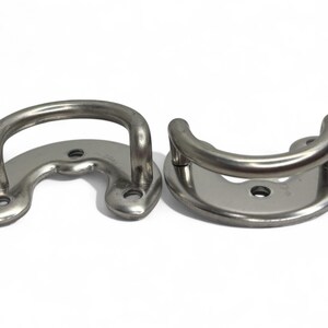 Peut inclure: Deux taquets en D en acier inoxydable. L'un a une poignée en forme de U et une base incurvée avec trois trous de montage. L'autre a une base arrondie avec une poignée en forme de U et deux trous de montage. Sur fond blanc.