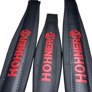May include: Three black Hohner accordion straps with red lettering and logo. The straps are made of a textured material and have a stitched edge. The Hohner logo is a red circle with a musical instrument design.