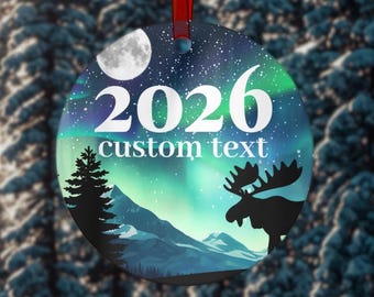 May include: Round Christmas ornament with a winter scene. The ornament features the year "2026" and the words "custom text" in white. A silhouette of a moose and a pine tree are visible. The background shows a night sky with the aurora borealis.