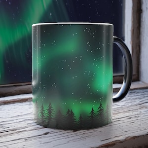 May include: A black ceramic mug with a green and white aurora borealis design and a silhouette of a forest at the bottom. The mug is sitting on a white wooden surface.