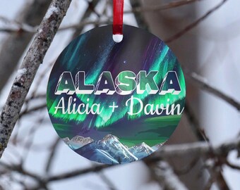 May include: Round Christmas ornament with a red ribbon, featuring the word "ALASKA" in white and green lettering. Below, the names "Alicia + Davin" are displayed. The design includes a vibrant aurora borealis and mountain range.