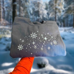 May include: A dark gray sweatshirt with a snowflake and star design. The design is white and light blue, with a gradient effect from dark gray to blue at the bottom. The sweatshirt is held up by a person wearing an orange sleeve.