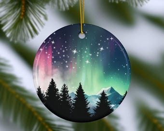May include: A round Christmas ornament featuring a colorful depiction of the Northern Lights. The design includes a starry sky, silhouetted trees, and mountains. The ornament is suspended by a gold string.